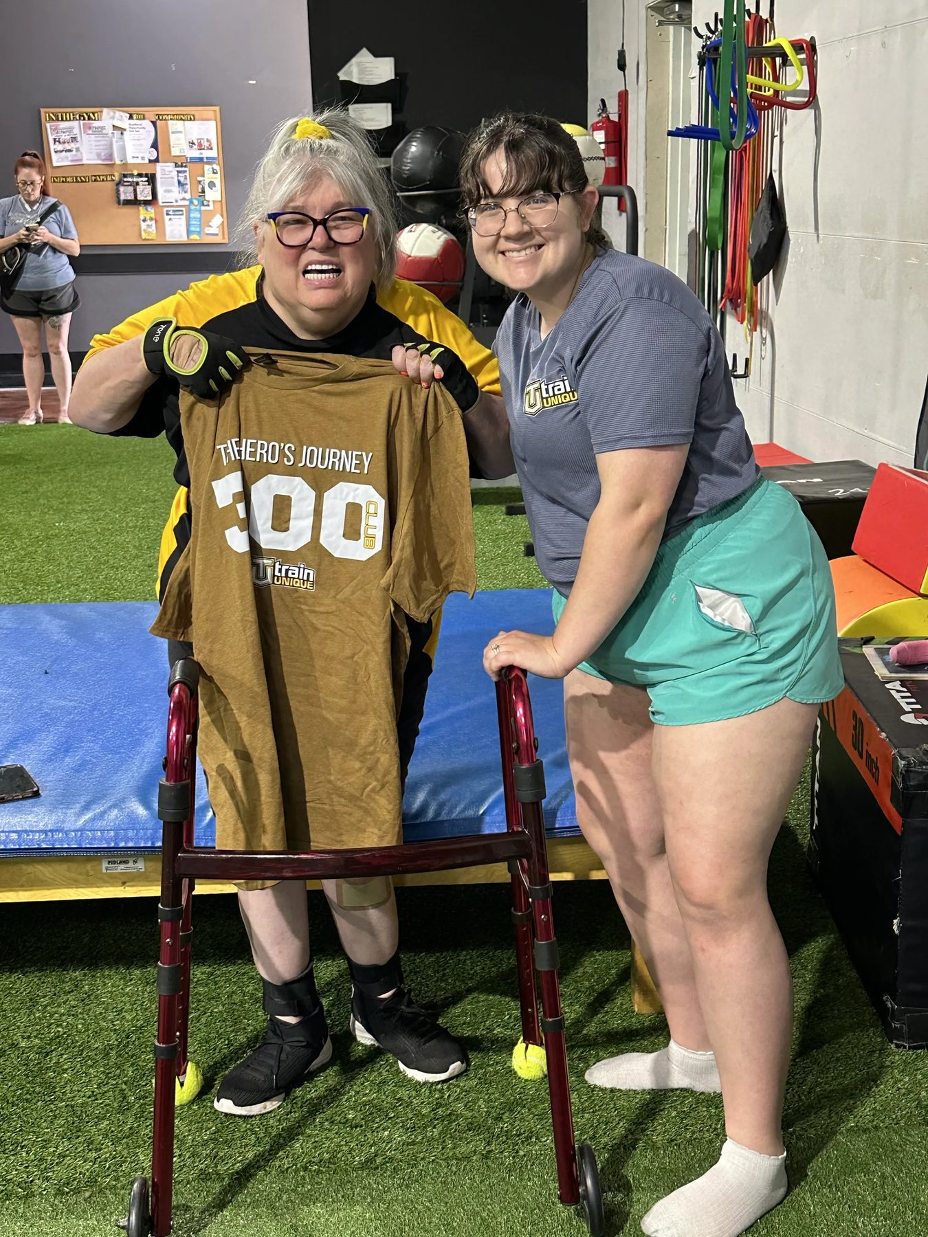 An athlete with a walker holds up her Hero's Journey 300 Club shirt beside a smiling coach in the Train Unique gym
