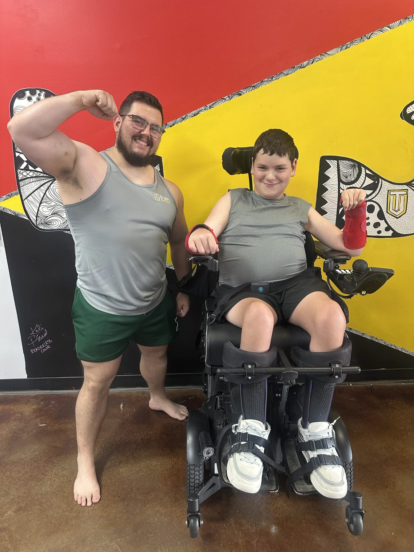 An athlete using a powerchair and a coach smile and flex together in front of the Train Unique gym mural
