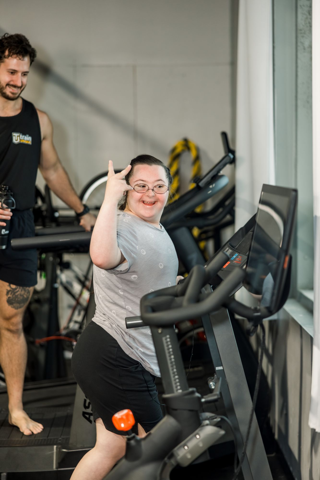An athlete on an exercise bike grins and makes a hand sign by her face while a coach behind her smiles