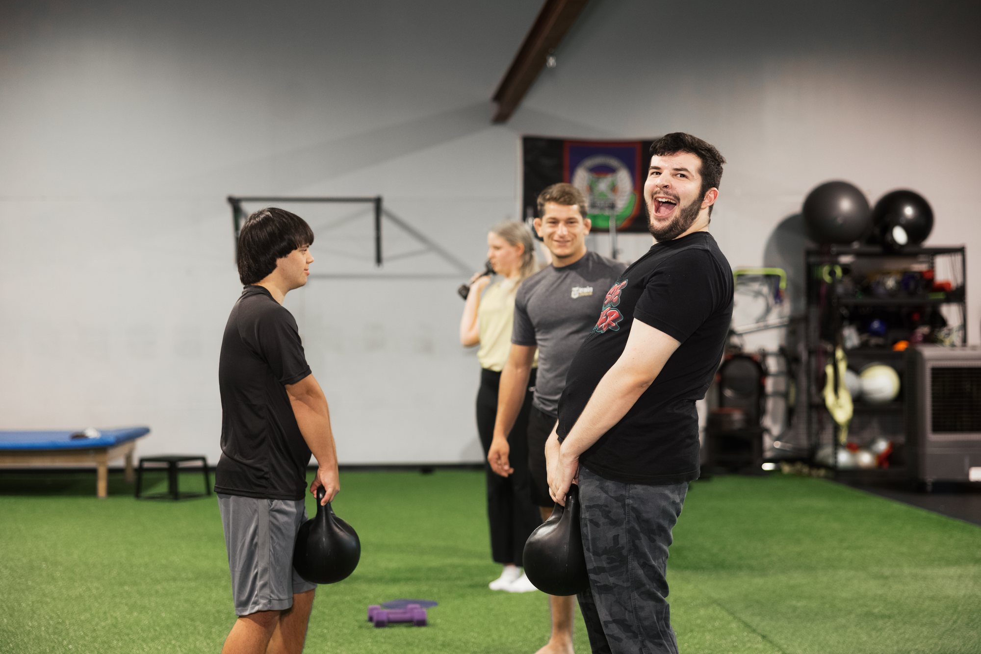 Two athletes hold kettlebells on the turf; one laughs with his mouth wide open while a coach in a Train Unique shirt smiles behind them