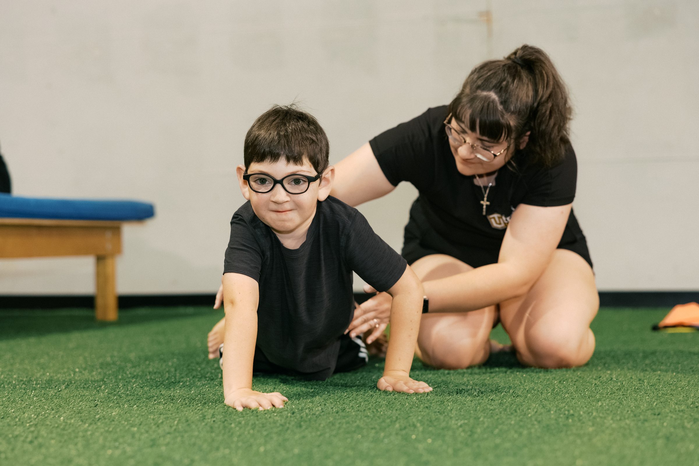 A young athlete in glasses grins in the middle of a bear crawl while his coach kneels beside him