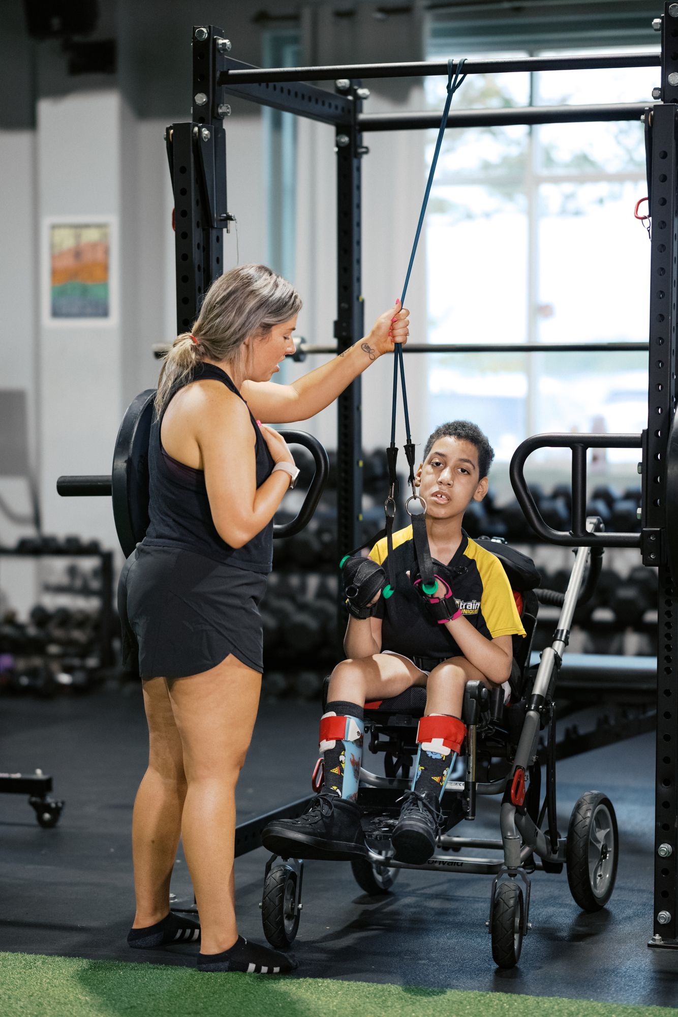 An athlete seated in his adaptive chair holds two cable handles while his coach adjusts the cable above him